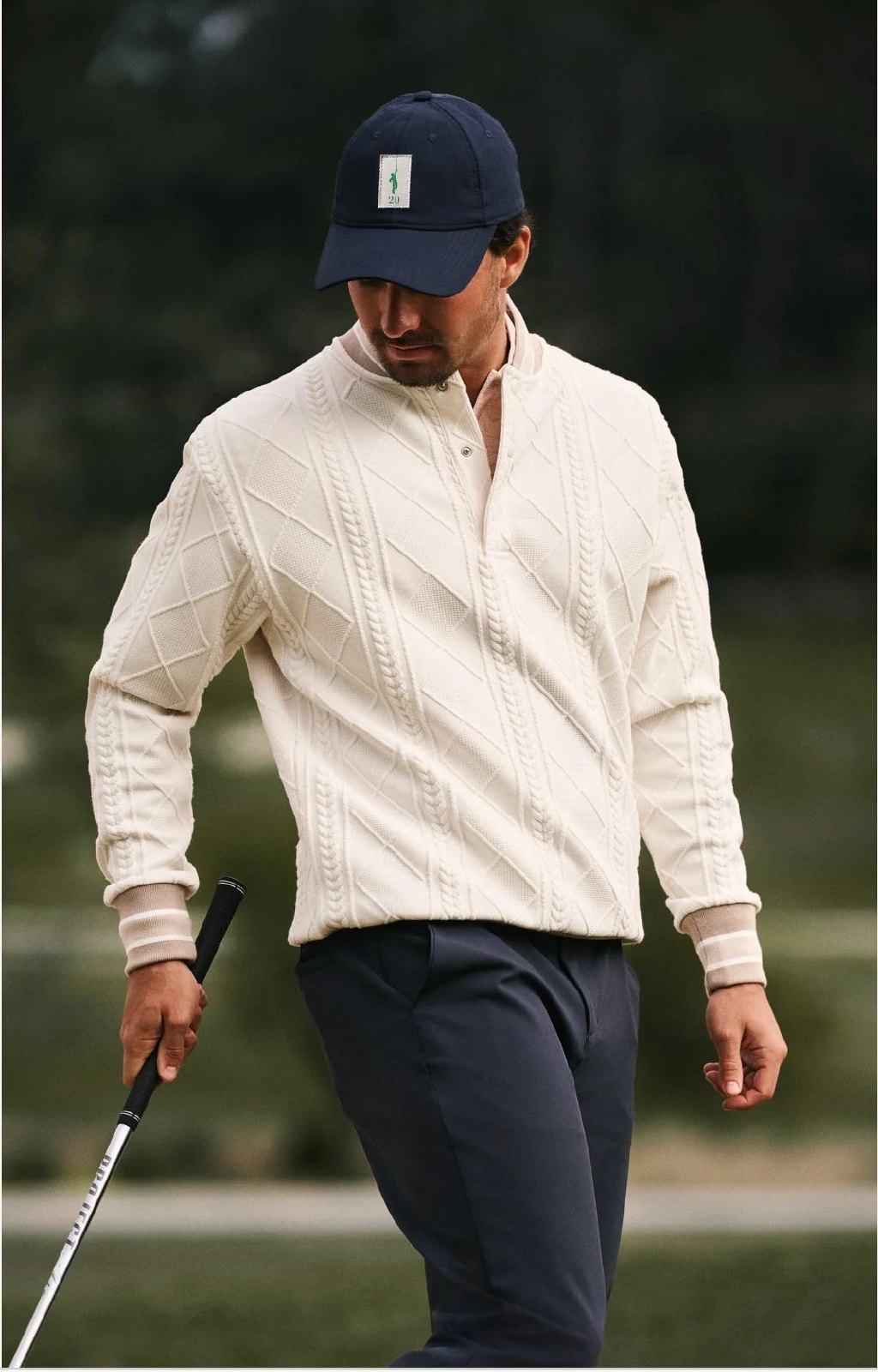Golfer in a natural cream cable-knit quarter-zip, navy cap and navy trousers.