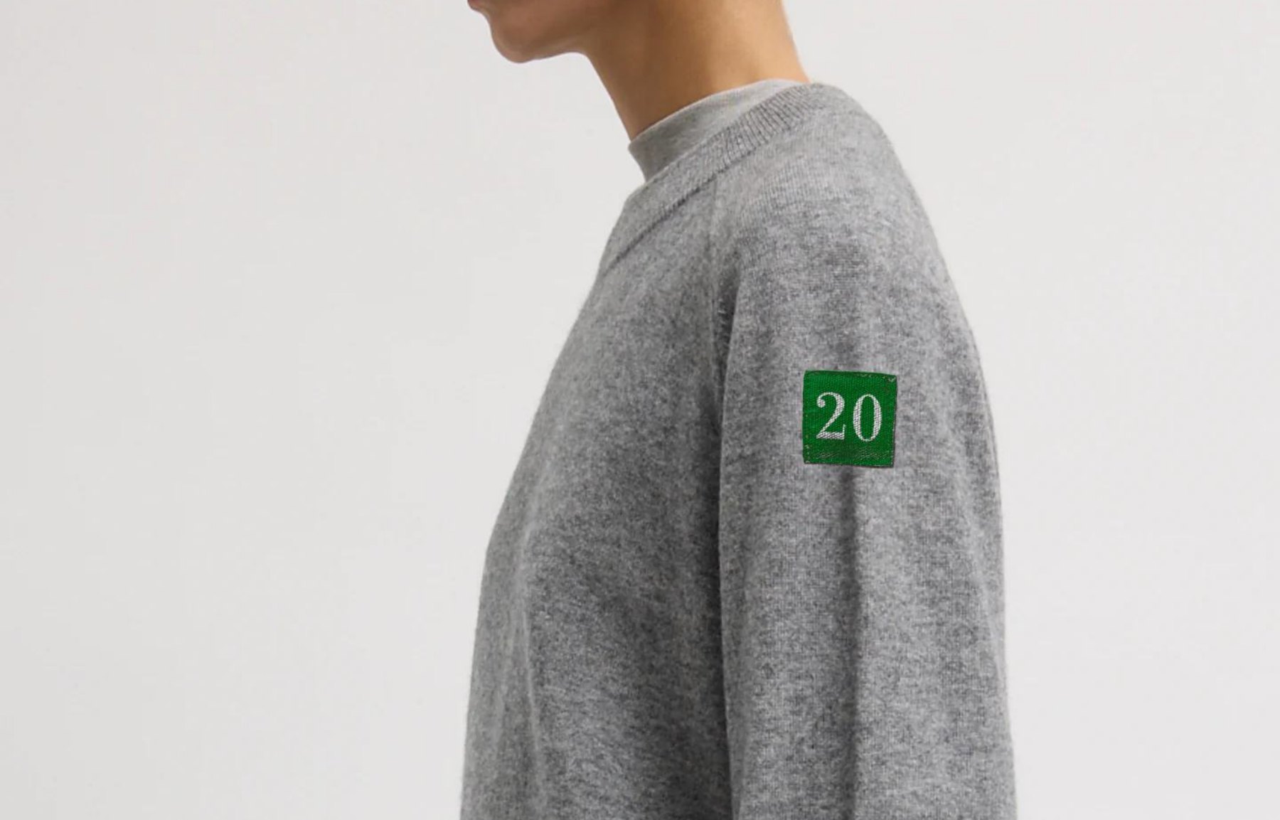Heather gray cashmere crewneck with a felted green 20 patch on the upper arm.