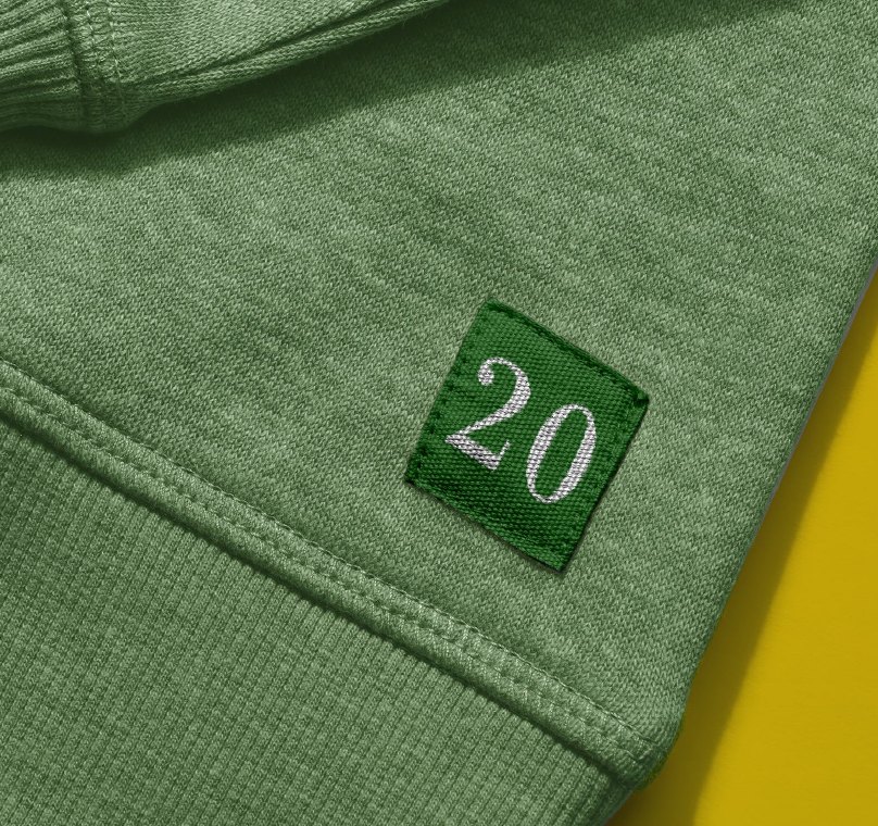 A square green felt 20 patch stitched to the hem of heather gray knitwear.