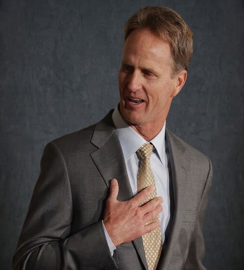 Jack Nicklaus II in a grey suit and gold patterned tie, hand on chest, against a dark studio backdrop.